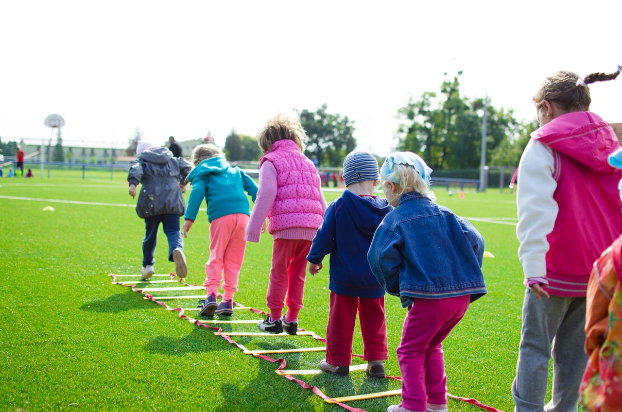 Home Children enjoy an outdoor activity on a grassy field, stepping over a ladder.