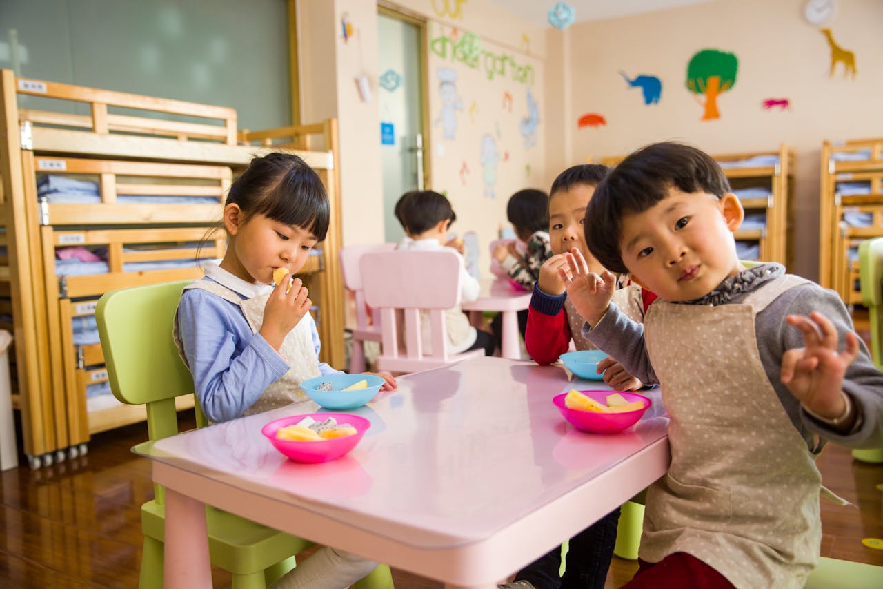 Home Kids seated around a table in a colorful classroom, eating snacks happily.